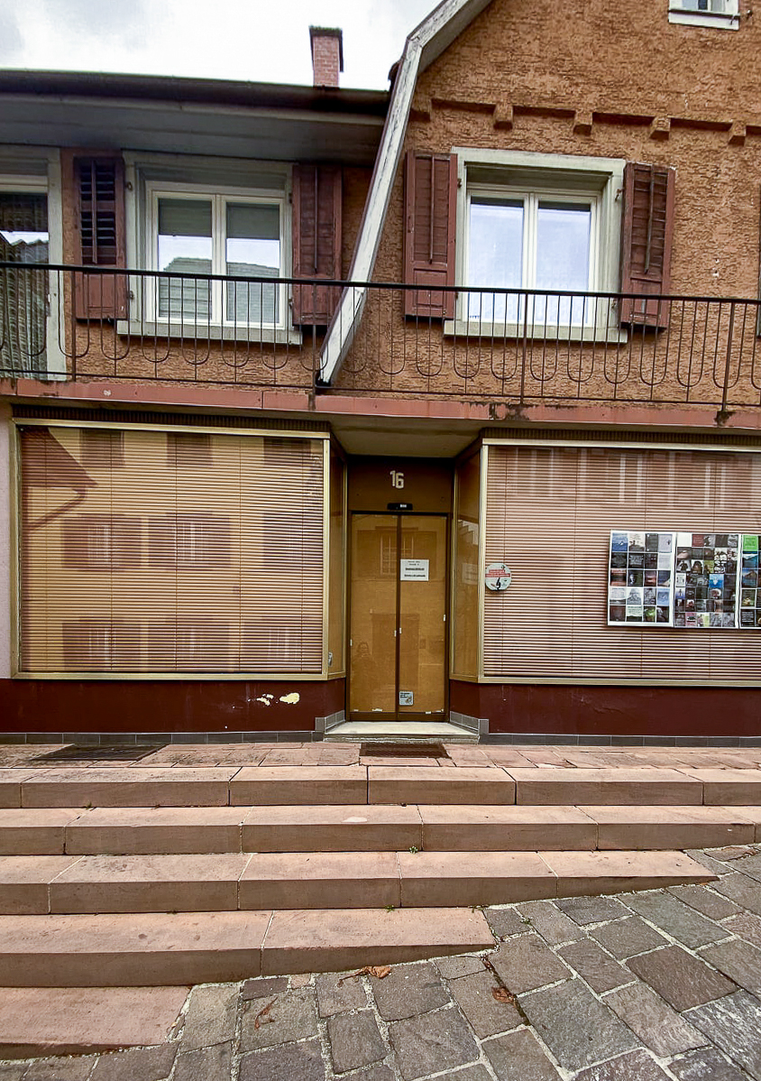 Ein zweistöckiges rötliches Gebäude in einer Altstadt. Eine Treppe führt zu einer Glastür die etwas nach innen gesetzt ist, zwischen den beiden Schaufenstern. Die Jalousie versperrt die Sicht nach innen in den Schaufenstern, innen gegen die Eingangtür sind Spanplatten gelehnt. Über dem Laden ist eine Wohnung.
