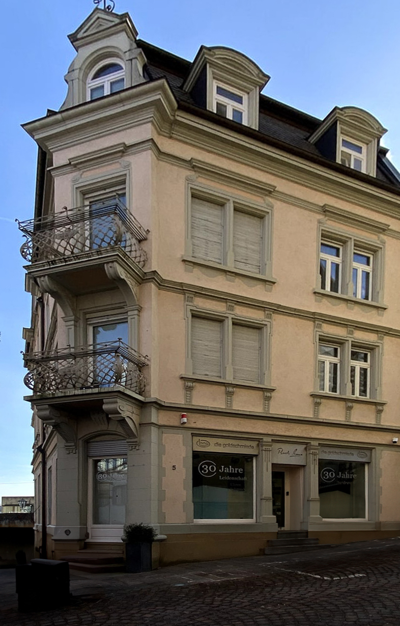 Ein hohes Eckgebäude. Im unteresten Stockwerk befindet sich ein Laden. Die Ecke ist wie abgeschnitten und dort befindet sihc der Eingang mit Stufen zum leerstehenden Geschäft.