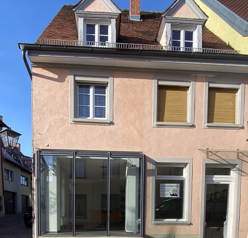 Ein rosafarbenes Eckhaus in einer Altstadt. Im untersten Stock ist ein Schaufenster, welches nach vorne raus steht. Rechts davon befindet sich der Eingang. Oben rechts ist eine Hausdekoration aus Metall mit einem Tier, dass eine Brezel vertilgt.